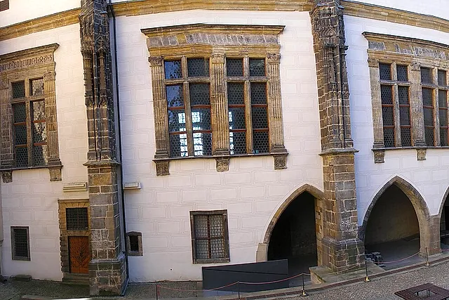 Eine weiß getünchte Mauer des Alten Königspalastes auf der Prager Burg, mit steingerahmten Fenstern, zwei Spitzbogenöffnungen und Eckstrebepfeilern aus Haustein über einem gepflasterten Hof.
