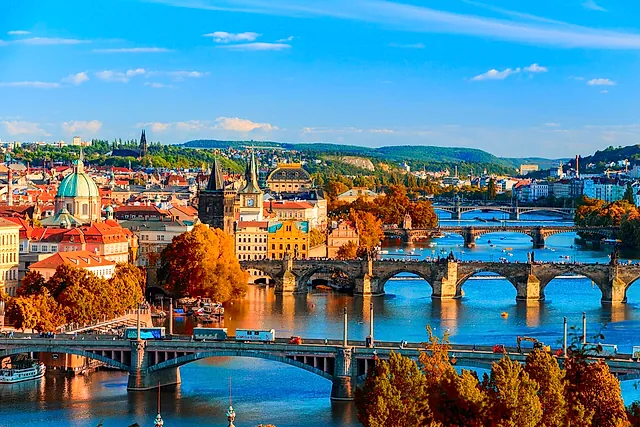 Panoramablick über Prags Dächer im Herbst, mit einer grünkuppeligen Kirche, orangefarbenen Bäumen und Bogenbrücken über den Fluss unter blauem Himmel.