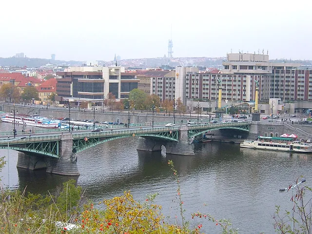 Blick aus dem Letná-Park auf die Čech-Brücke, eine grüne Stahlbrücke über die Moldau, mit weißen Booten am Ufer und Bürobauten dahinter.