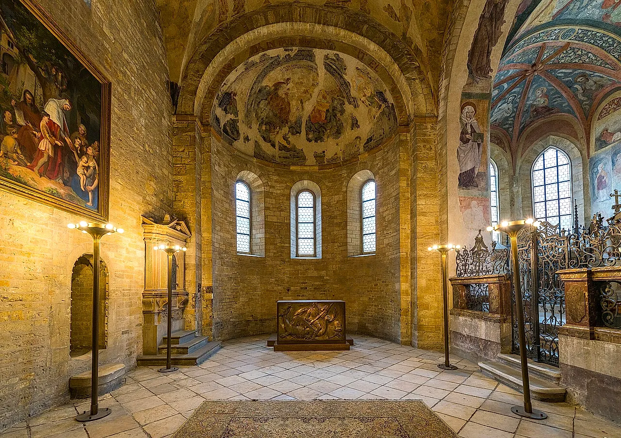 Interno della Basilica di San Giorgio al Castello di Praga, con l’abside medievale, affreschi sbiaditi nella volta, strette finestre ad arco e una cassa di metallo con un rilievo davanti all’abside.