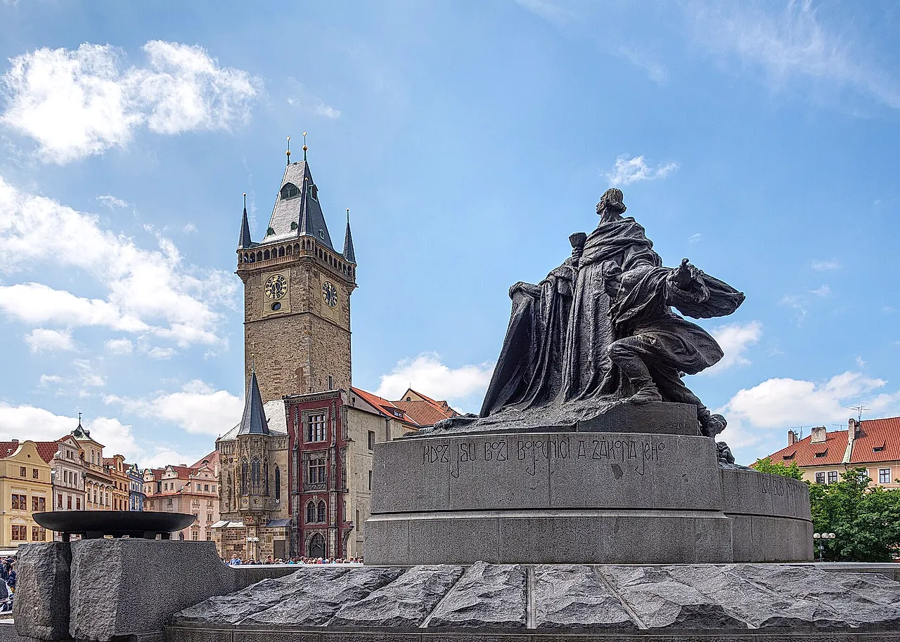 Das Jan-Hus-Denkmal auf dem Altstädter Ring, eine Bronzefigur auf einem Granitsockel, dahinter erhebt sich der Altstädter Rathausturm unter blauem Himmel.