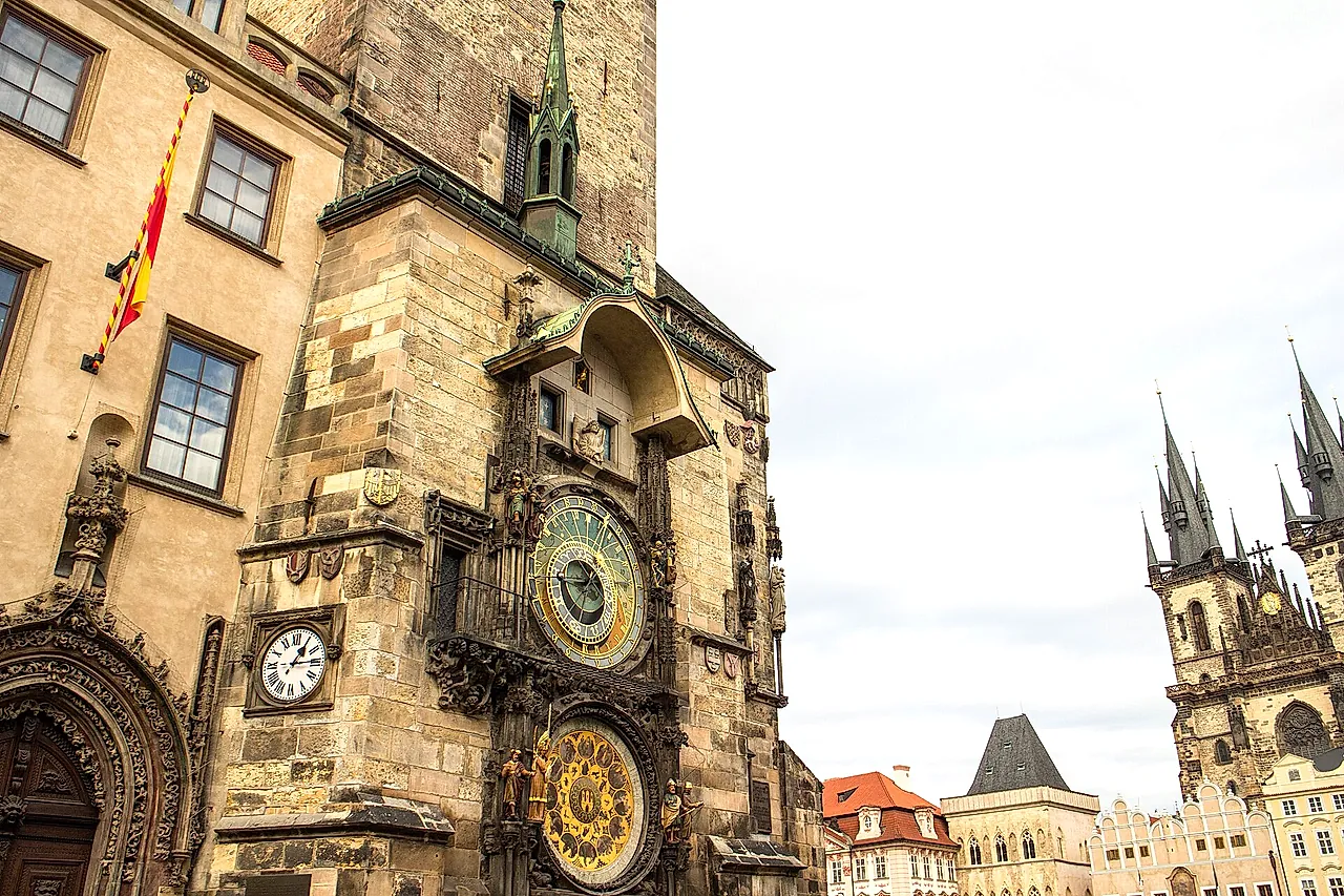 Die Astronomische Uhr am Altstädter Rathausturm, von der Seite gesehen, mit ihrem blauen astronomischen Zifferblatt und dem Kalenderblatt darunter und den Türmen der Teynkirche dahinter.