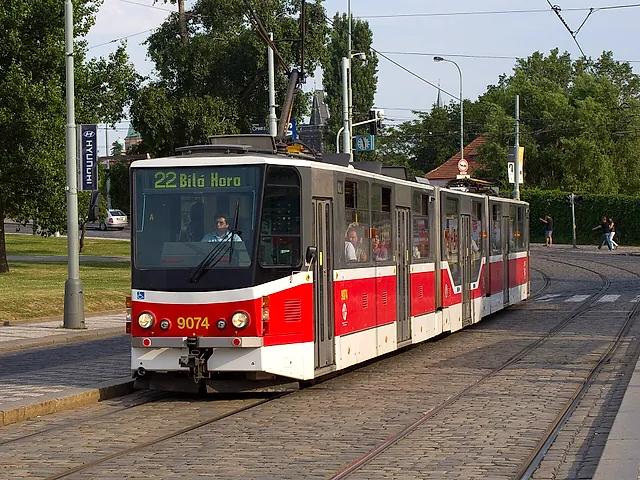 Eine rot-weiße Straßenbahn der Linie 22 mit der Zielanzeige Bílá Hora fährt auf Gleisen im Kopfsteinpflaster neben einem Grasstreifen und hohen Bäumen.