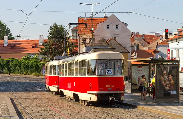 Die rot-cremefarbene Straßenbahn 8013 der Linie 23 wartet an einer Haltestelle, rechts ein Glasunterstand und dahinter orangebedachte Häuser unter blassem Himmel.