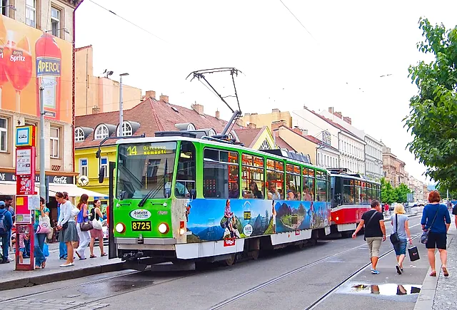 Tram number 14 at a Prague stop: a green-and-white tram with a red tram behind, passengers on the pavement and an Aperol billboard on a building.
