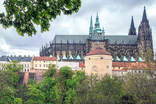 Der Veitsdom von der Nordseite, mit gotischen Strebebögen und zwei Türmen über einem runden Steinturm mit rotem Dach, unter bewölktem Himmel.