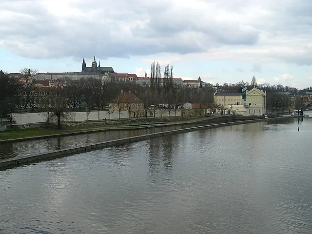 Die Moldau fließt an einer Ufermauer und Uferbäumen vorbei, mit der Prager Burg und ihren Domtürmen auf dem Hügel dahinter unter bewölktem Himmel.