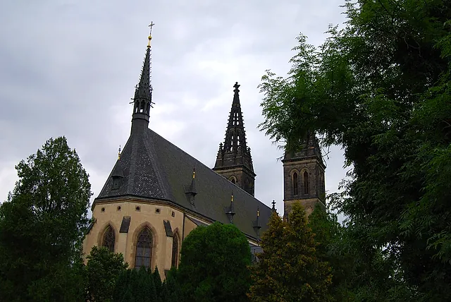 Eine gotische Kirche mit steilem grauem Dach und zwei spitzen Türmen, die über grünen Bäumen und einer niedrigeren Steinkapelle mit Bogenfenstern aufragt, unter bewölktem Himmel.