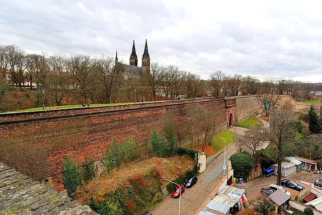 Rote Backsteinmauer der Festung Vyšehrad, die sich auf ein Tor zubiegt, mit den Zwillingstürmen der Basilika über den Bäumen und parkenden Autos auf der Straße darunter.