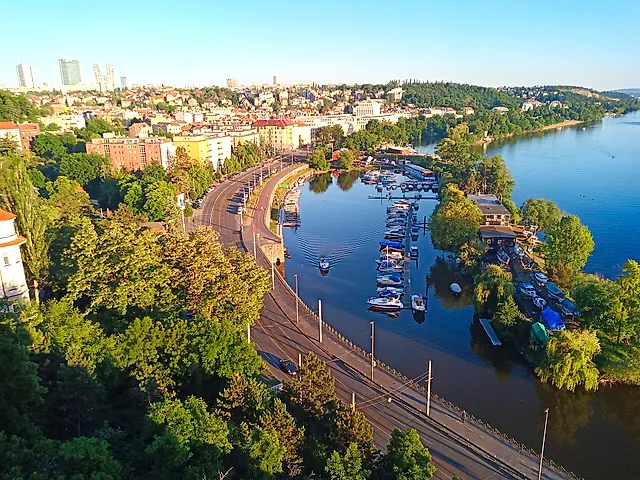 Luftaufnahme eines Moldau-Yachthafens mit festgemachten Booten, einer baumgesäumten Uferstraße und Häuserblöcken und bewaldeten Hügeln dahinter unter klarem blauem Himmel.