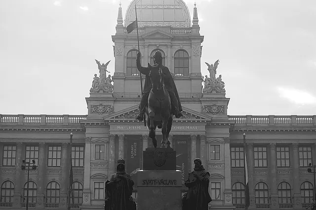 Schwarz-weiße Ansicht des Nationalmuseums in Prag, mit seiner verglasten Kuppel, Skulpturen auf der Dachlinie und dem Reiterdenkmal des heiligen Wenzel davor, unter grauem Himmel.