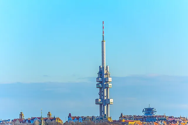 Der Fernsehturm Žižkov aus der Ferne vor blassblauem Himmel, seine Betonkabinen und der hohe Mast ragen über die Dächer der Stadt.