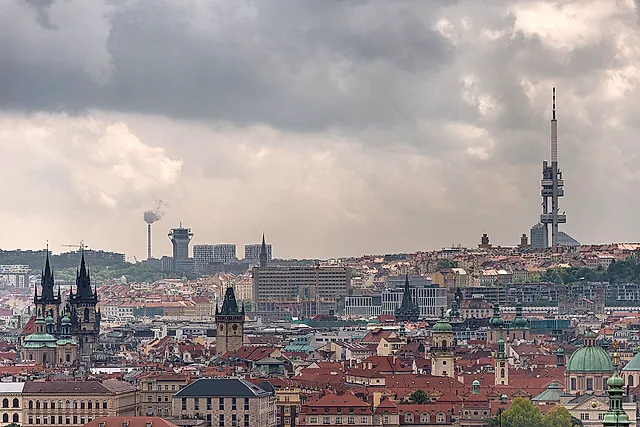 Der Fernsehturm Žižkov rechts in einem Blick über Prags rote Dächer, mit den Zwillingstürmen der Teynkirche links unter schwerem grauem Himmel.