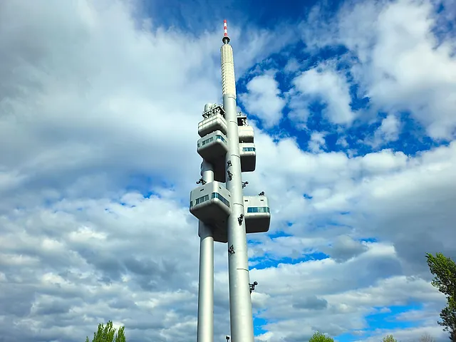 Žižkov Television Tower photographed from below against a blue sky with white clouds, its pods and mast rising above the trees, with small sculptures on the shaft.
