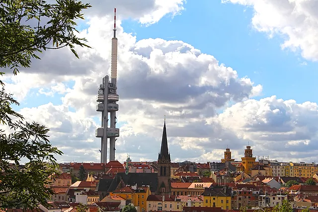 Žižkov Television Tower seen across the city rooftops under a blue sky with large white clouds, its pods and red-tipped mast rising above the rooftops and a church spire.